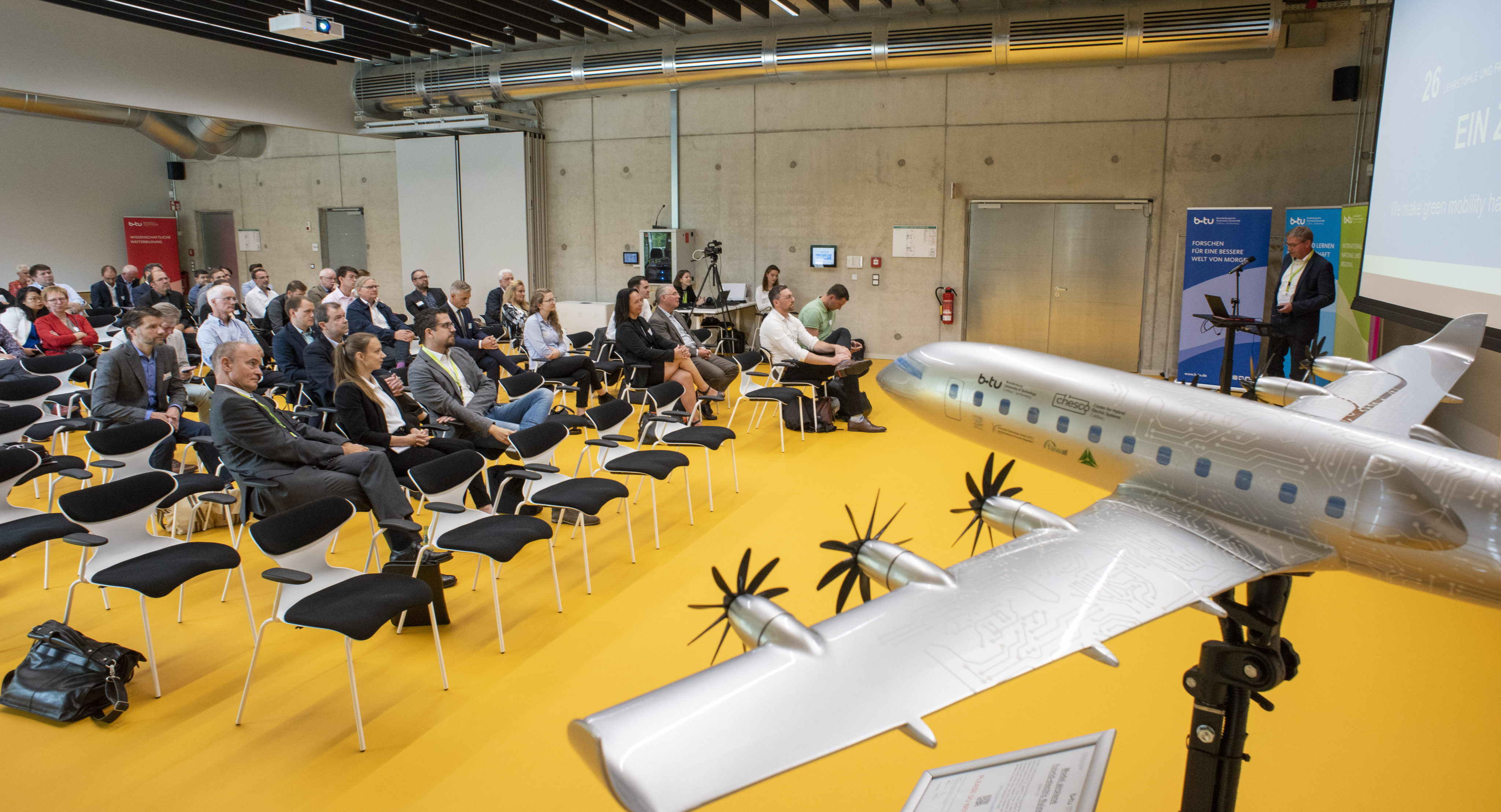 BTU Veranstaltung Blick auf Plenum/ Podium mit Modellflugzeug im Vordergrund